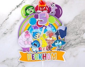 May include: A colorful cake topper featuring characters from the movie Inside Out. The topper includes Joy, Sadness, Anger, Fear, Disgust, and Bing Bong. The topper also includes a rainbow and the number 5. The topper is personalized with the name "BECKHAM".