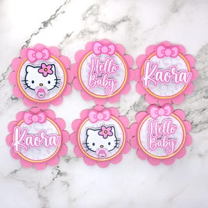 Könnte beinhalten: Sechs rosa blütenförmige Dekorationen mit Hello Kitty-Motiv. Jede Dekoration hat einen rosa gewellten Rand, eine Schleife und eine kreisförmige Mitte. Einige zeigen den Text "Hello Baby" oder den Namen "Kaora". Andere zeigen Hello Kitty mit einem Schnuller.