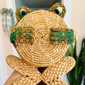 May include: A woven wicker frog with green painted details and large black eyes. The frog is sitting on a hand.