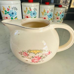May include: A vintage cream-colored pitcher with a floral design featuring pink roses and green leaves. The pitcher has a wide mouth and a handle.