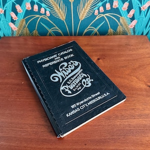 May include: A vintage black book titled "Physicians' Catalog and Reference Book" from Wise's Pharmacy Co. The book is on a wooden surface, with text including the pharmacy's name and address in Kansas City, Missouri, USA.