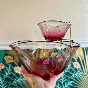 May include: A two-tiered serving set featuring a large, clear glass bowl and a smaller bowl with a pink ombre effect. The bowls have a wavy rim and are held by a gold-colored metal stand. The set is ideal for serving snacks or dips.