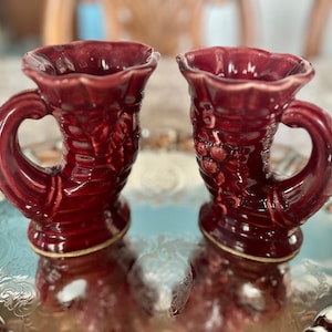 May include: Two small, burgundy ceramic vases with a floral design. The vases have a handle and a wide opening at the top.