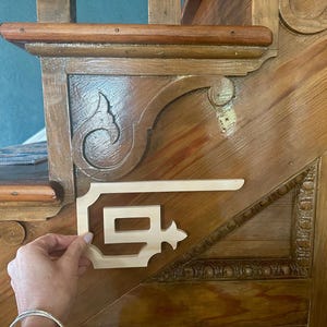 May include: A wooden stair detail with ornate carvings and a light-colored wooden decorative piece being held in place. The stair is made of stained wood with a natural finish, showcasing the wood grain.