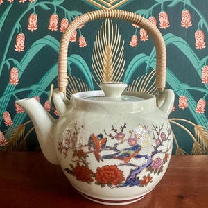 May include: A cream-colored ceramic teapot with a crackle glaze finish, featuring a curved spout and a light brown woven rattan handle. The body is decorated with a colorful painted scene of birds perched on flowering branches with red and pink blossoms.