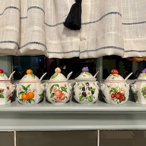 May include: A collection of six white ceramic jars with lids and spoons, each decorated with a different fruit design. The fruits include strawberries, oranges, peaches, blackberries, cherries, and grapes. The jars are arranged on a shelf.