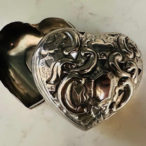 May include: A silver heart-shaped trinket box with an ornate, detailed design. The box is open, revealing the interior.