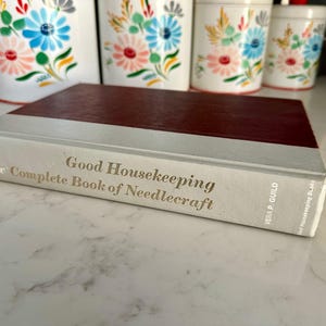 May include: A hardcover book titled "Good Housekeeping New Complete Book of Needlecraft" with a burgundy cover and white spine. The book is lying on a white surface, with floral canisters in the background.