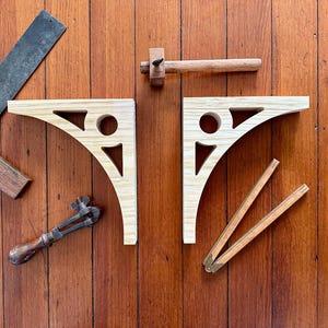 May include: Overhead shot of woodworking tools on a wooden surface. Includes two light-colored wooden shelf brackets with decorative cutouts, a metal and wood square, a small wooden mallet, a metal clamp, and a wooden measuring tool.