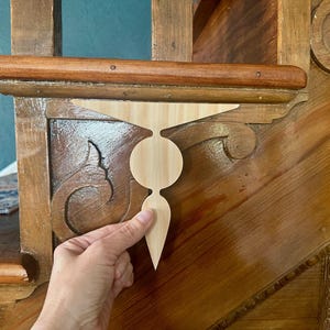 May include: A light-colored wooden decorative bracket with a unique shape, being held up against a wooden staircase. The bracket has a circular center and a pointed bottom, designed to be an accent piece.
