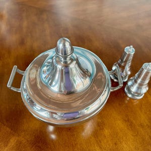 May include: A silver serving dish with a lid and two matching salt and pepper shakers. The dish has a round, fluted design and a handle on each side. The shakers are tall and slender with a fluted design.