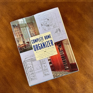 May include: The Complete Home Organizer book cover features a blue, white, and black design with illustrations of different rooms and storage solutions. The title "The Complete Home Organizer" is in bold, black text with the author's name, Maxine Ordesky, in smaller black text below. The book cover also includes the text "A guide to functional storage space for all the rooms in your home."
