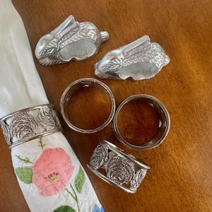 May include: Four silver napkin rings, two are shaped like rabbits and two are ornate with a floral design. The rings are on a white napkin with pink flowers.