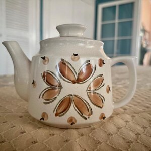 May include: A white ceramic teapot with a brown floral design. The teapot has a rounded body and a spout on the side. The handle is on the opposite side of the spout.
