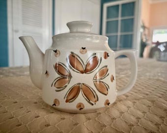 May include: A white ceramic teapot with a brown floral design. The teapot has a rounded body and a spout on the side. The handle is on the opposite side of the spout.