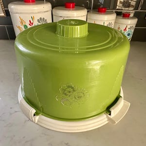 May include: A green cake dome with a white base and a floral design. The dome has a hinged lid and a white knob on top.