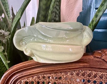 May include: A light green ceramic planter with a unique, abstract design. The planter has a smooth, glossy finish and is shaped like a wave.
