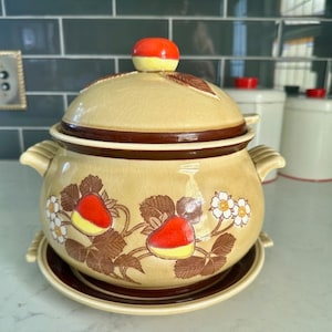 May include: A vintage ceramic serving bowl with a lid. The bowl is decorated with a brown, yellow, and red strawberry pattern. The lid has a red and yellow strawberry on top.
