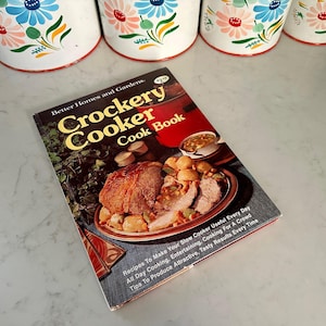 May include: A vintage "Crockery Cooker Cook Book" by Better Homes and Gardens. The cookbook features a photo of a roast with vegetables. White and floral-decorated containers are in the background.