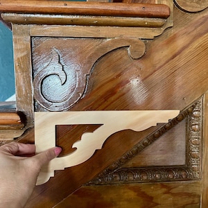 May include: A light-colored wooden corbel held against a dark wood surface. The corbel has a decorative, ornate design. The background features a wooden staircase with carved details. The corbel is likely for home decor or woodworking projects.