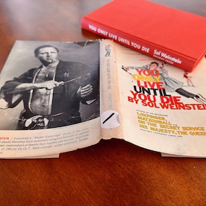 May include: A book titled "You Only Live Until You Die" by Sol Weinstein. The cover features a man in a red jacket and white pants jumping in the air. The book is open to a page with a black and white photo of a man in a black kimono holding a knife.