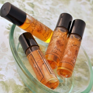 Puede incluir: Cuatro peque&ntilde;os frascos de perfume de vidrio con tapas negras y l&iacute;quido &aacute;mbar est&aacute;n dispuestos en un plato de vidrio transparente. Las botellas est&aacute;n etiquetadas como "Sweet Sally's Patchouli Perfume Oil". El plato est&aacute; sobre una superficie con estampado floral.