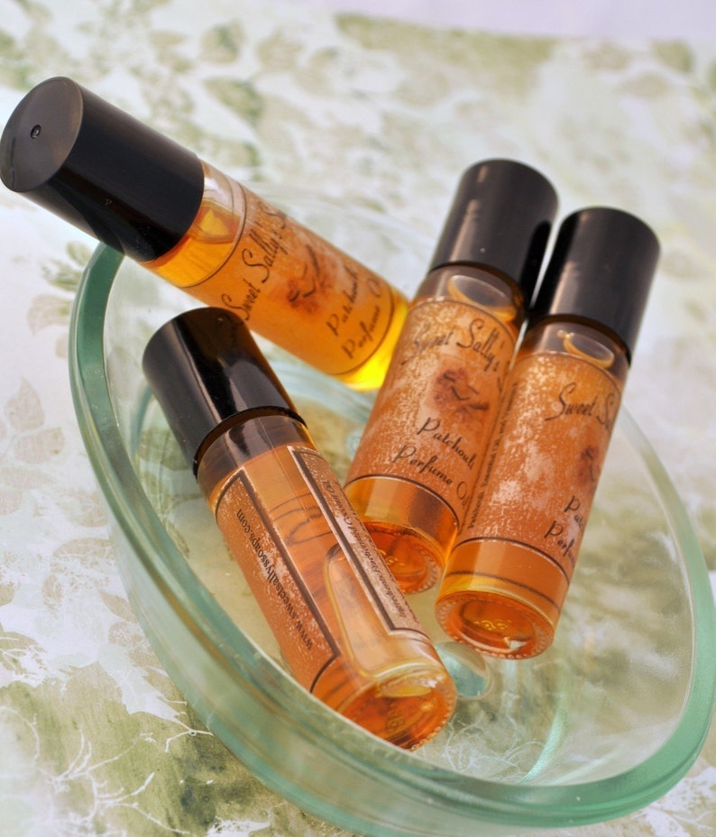 Puede incluir: Cuatro peque&ntilde;os frascos de perfume de vidrio con tapas negras y l&iacute;quido &aacute;mbar est&aacute;n dispuestos en un plato de vidrio transparente. Las botellas est&aacute;n etiquetadas como "Sweet Sally's Patchouli Perfume Oil". El plato est&aacute; sobre una superficie con estampado floral.