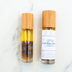 May include: Two glass essential oil roller bottles with wooden caps. The bottles contain a yellow liquid and dark brown stones. One bottle has a label that reads "Third Eye Cho" and "Essential Oil Blend".