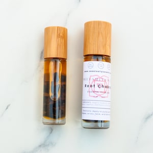 May include: Two glass essential oil roller bottles with wooden caps. One bottle has a label that reads "Root Chakra Essential Oil Blend" and "www.sweetsallysscents". The bottles contain a dark liquid and are set against a white marble background.