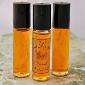 Puede incluir: Tres frascos de perfume roll-on de vidrio transparente con tapas negras. Los frascos contienen un l&iacute;quido dorado. Un frasco tiene una etiqueta que dice "Sweet Sally's Soap Patchouli Perfume Oil."