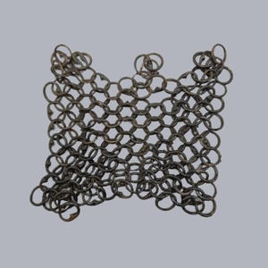 Può includere: Primo piano di una sezione di armatura a maglia di catena. Gli anelli metallici sono interconnessi in un motivo esagonale. La cotta di maglia è di un colore metallico scuro e invecchiato, che suggerisce che sia antica o vintage.