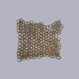 Può includere: Un frammento di armatura a maglia di catena antica. Gli anelli metallici sono interconnessi in un motivo denso, mostrando segni di età e usura. La cotta di maglia ha una forma quadrata con un bordo leggermente irregolare.