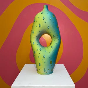 Könnte beinhalten: Eine grüne und blaue Keramikvase mit einem Loch in der Mitte und einer strukturierten Oberfläche, die an Regentropfen erinnert. Die Vase steht auf einem weißen Sockel vor einem rosa und orangenen Hintergrund.