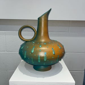 May include: A decorative ceramic vase with a unique design. The vase features a bulbous body with a handle and a spout. It is painted in shades of teal and gold, with teal teardrop accents. The vase is displayed on a white pedestal.