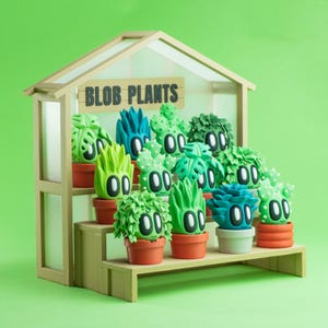 May include: A wooden greenhouse display with the text "BLOB PLANTS" features various green and blue blob-shaped plant sculptures in terracotta pots. The plants have cartoonish faces with black eyes and white highlights, arranged on tiered shelves.