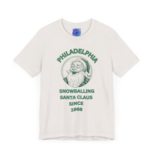 May include: White t-shirt with a green graphic of Santa Claus laughing and the text "Philadelphia Snowballing Santa Claus Since 1968."