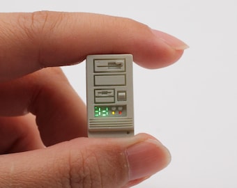 Puede incluir: Una torre de ordenador blanca en miniatura con una pantalla digital verde que muestra "3:21" y luces rojas y amarillas. La torre tiene una serie de ranuras y botones.