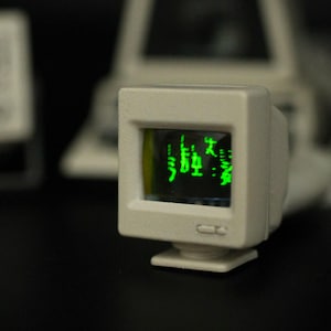 Könnte beinhalten: Ein Miniatur-Computerbildschirm in Off-White mit einem kleinen Bildschirm, der grünen Text anzeigt. Der Monitor hat eine quadratische Form und einen kleinen Sockel. Weitere Computerkomponenten sind im Hintergrund sichtbar.