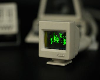 May include: A miniature beige computer monitor with a square screen displaying bright green Japanese characters. The device features a small rectangular base and a compact, boxy frame, capturing the aesthetic of vintage desktop technology.