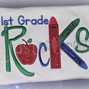 May include: A white t-shirt with the text "1st Grade Rocks" in green, blue, and red glitter. The text is surrounded by a red apple, a blue crayon, and a green crayon.