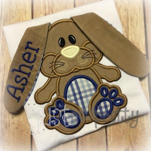 Può includere: Un'applicazione di coniglio marrone con tessuto a quadri blu e bianchi su una maglietta bianca. Il coniglio ha lunghe orecchie marroni con il nome "Asher" ricamato su un orecchio.