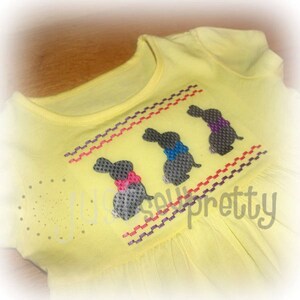May include: A yellow children's dress with three embroidered gray bunnies wearing colorful bows. The bunnies are arranged in a row and are stitched with a purple, red, and white pattern.