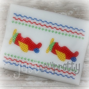 May include: Two red, yellow, and blue airplanes with propeller embroidery on a white fabric background with a repeating pattern of blue, red, and green lines.