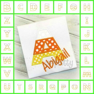 May include: A white t-shirt with an orange and yellow candy corn letter A and the name Abigail embroidered on it. The letter A is surrounded by a white outline. The name is in a cursive font. The t-shirt is surrounded by a grid of letters of the alphabet in a gold and white striped pattern.