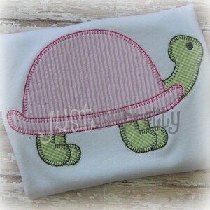 Puede incluir: Una aplicación de tortuga rosa y verde con un caparazón a rayas. La tortuga está cosida sobre una tela azul claro.