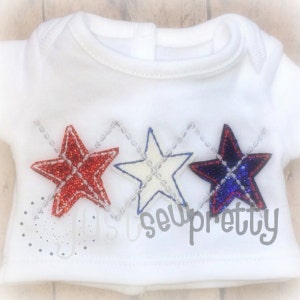 May include: A white baby onesie with three embroidered stars in red, white, and blue. The stars are arranged in a diagonal pattern and have a diamond pattern inside.
