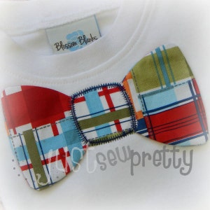 May include: A white shirt with a red, blue, and green plaid bow tie. The bow tie has a stitched edge and the text "Blossom Blanks" is on the shirt.