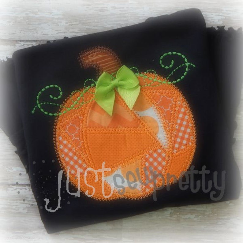 Puede incluir: Una tela negra con un dise&ntilde;o de calabaza de patchwork. La calabaza es naranja con hojas verdes y un lazo verde. La tela tiene el texto "Just Sew Pretty" bordado.