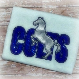 Puede incluir: Una camiseta azul claro con la palabra "COLTS" en letras azules llenas de purpurina. Un caballo plateado en posición de encabritado está centrado encima de la palabra. La camiseta es probablemente para un equipo deportivo o ropa de aficionado.