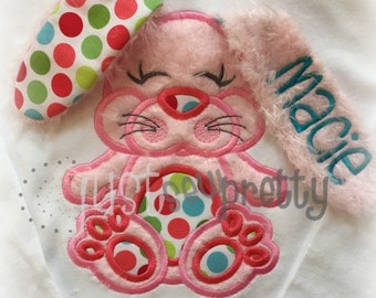 Puede incluir: Aplique de conejito rosa con un patrón de lunares y una cola esponjosa. El conejito tiene los ojos cerrados y una nariz rosa. La cola está bordada con el nombre "Maddie".